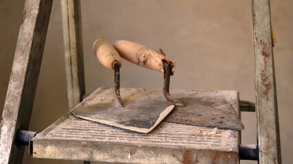 two old shabby trowels on the steps of a metal stepladder against the background of light plastered walls as a construction composition in a pause of renovation work