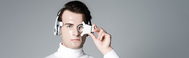 Cyborg in headphones looking at camera while holding digital eye lens isolated on grey, banner
