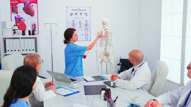 Young female assistent pointing on skeleton in front of medical surgeons in conference room. Clinic expert therapist talking with colleagues about disease, medicine professional