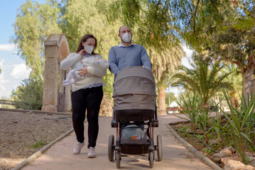 Happy couple walking in the park with stroller. They wear medical masks to protect them from the virus.