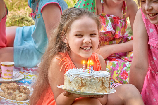 Beautiful happy little girl holding a cake in a large group of friends. Celebrate your birthday in nature. Picnic in the Park in summer.