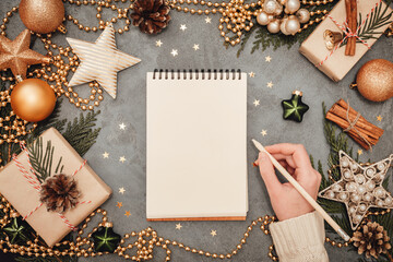 Notepad and writing female hand with holiday decorations in green and golden colors on grey concrete backdrop, copyspace. Christmas and New Year planning flatlay concept.