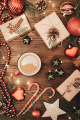 Christmas and New Year rustic traditional composition. Hot coffee cup. Festively decorated cedar branches on dark brown old wooden board. Gift boxes, balls, stars and cane lollypops.