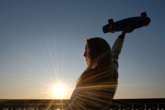 Silhouette of the extatic woman with skateboard in her raised in sunset sky hand, side view, vintage color.