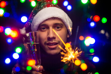 Young guy with a glass of champagne and a sparkler. Christmas garland. Blurred foreground. New Year's bokeh. Light of New Year's lights. Happy New Year.