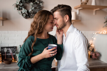 Husband and wife, alike, have fun together in a Scandinavian-style kitchen. The kitchen is decorated before Christmas. The couple hugs.