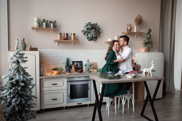 Fototapeta na wymiar Husband and wife, alike, have fun together in a Scandinavian-style kitchen. The kitchen is decorated before Christmas. The couple hugs.