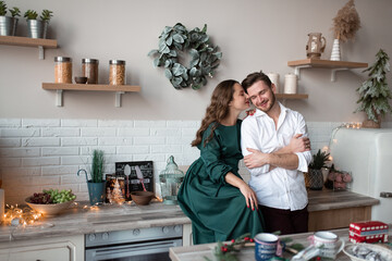 Husband and wife, alike, have fun together in a Scandinavian-style kitchen. The kitchen is decorated before Christmas. The wife kisses her beloved on the cheek.