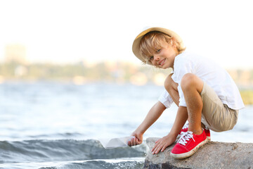 Cute little boy with paper ship at river, space for text. Child spending time in nature