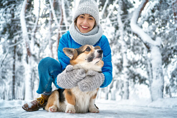 Winter smiling woman in warm hat and scarf hugging with her pet Welsh Corgi dog, having fun in snow at winter park