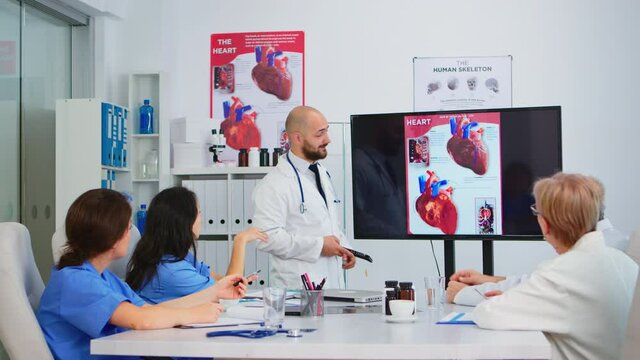 Experienced man doctor analysing heart issues image together with cvalified colleagues in meeting room, pointing on monitor. Doctors discussing diagnosis about treatment of patients