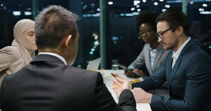 Rear of man boss speak with colleagues on team meeting in international company in modern office in evening Caucasian, African American and Arabic workers talking about business and tapping on tablets