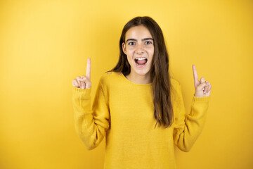 Pretty girl wearing a yellow sweater standing over isolated yellow background amazed and surprised looking at the camera and pointing up with fingers and raised arms