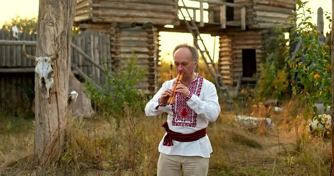 Man in a folk Slavic-Ukrainian costume plays a double pipe in a village courtyard. Traditions and rituals of the Slavic peoples. 4k, ProRes