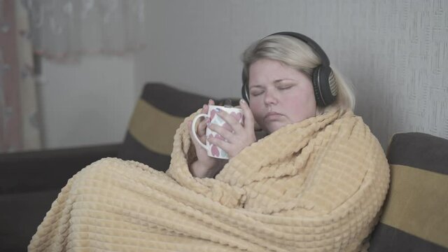 Fat woman wrapped in a blanket drinks tea and sings while listening to music on headphones.