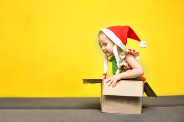 little girl in Santa's hat and an elf assistant costume sits in Cardboard Box. Concept of receiving or shipping Christmas parcel at home. delivery service celebrates Christmas.
