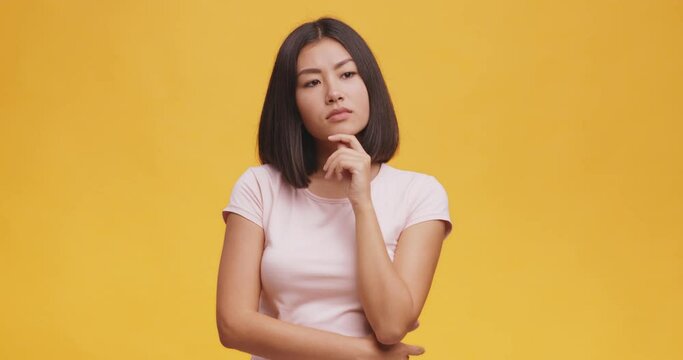 Young thoughtful asian woman pondering about something, touching her chin, orange studio background