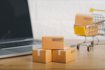 Fototapeta premium Brown paper boxs and cart with laptop on wood table in office background.Easy shopping with finger tips for consumers.Online shopping and delivery service concept.