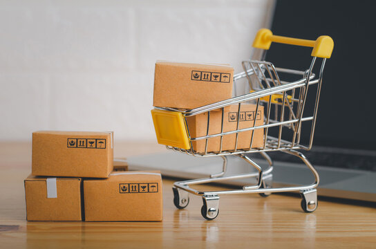 Brown paper boxs in shopping cart with laptop on wood table in office background.Easy shopping with finger tips for consumers.Online shopping and delivery service concept.