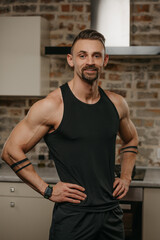 Obraz premium A smiling muscular man with a beard is posing in his apartment after a workout. The athletic guy is demonstrating his sporty physique at home. A smartwatch is on the wrist.