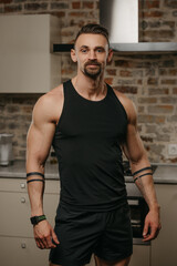 Obraz premium A kind muscular man with a beard is posing in his apartment after a workout. An athletic guy with tattoos on his arms is demonstrating his sporty physique at home. A smartwatch is on the wrist.