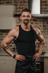 Fototapeta na wymiar A smiling muscular man with a beard is posing with his hands on a waist in his apartment after a workout. An athletic guy with tattoos on his arms is demonstrating his sporty physique at home.