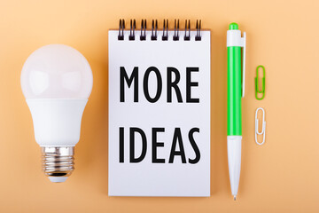 Text MORE IDEAS on a white piece of paper in notebook next to a light bulb and a green pen. Imagination concept.