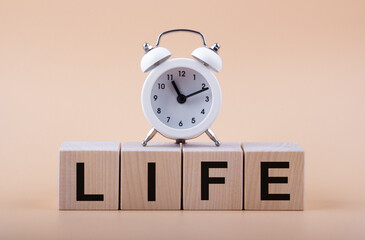 Text LIFE on wooden blocks and white alarm clock on a light peach background. The concept of evolution in time.