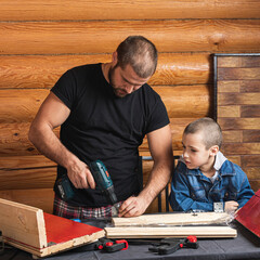 Father and son together make a wooden birdhouse in the workshop. Cheerful father with a little boy drilling a wooden plank using screwdriver  on a wooden table in the workshop