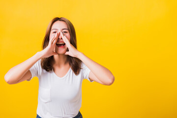 Asian Thai happy portrait beautiful cute young woman teen standing big shout out with hands next mouth giving excited positive looking to camera isolated, on yellow background with copy space