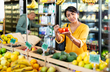 Interested Latina choosing ripe red tomatoes while shopping in vegetable department of grocery store