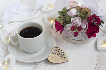 cup of coffee, carved wooden heart, a bouquet of small red and pink roses with eucalyptus leaves on a white wooden background
