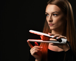 Young pensive woman with long silky straight hair in black bodysuit holds demonstrates hair straightener and looks aside at copy space over dark background. Haircare, beauty, wellness concept