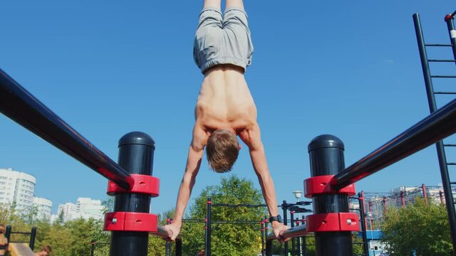 A man does a street workout at a sports ground. A sportsman does a handstand on parallel bars against an urban landscape