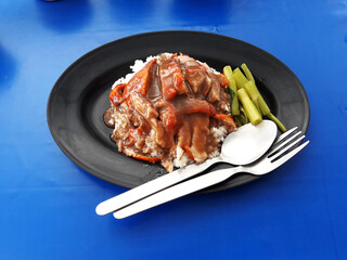 Roasted duck topping on rice with gravy sauce and green vegetable in black color dish on blue table, Thailand street food