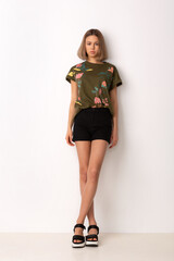young girl with skillful makeup in black shorts and a sweater with a print of flowers posing standing against a white wall