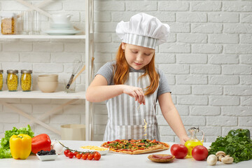 little girl in chef hat and an apron cooking pizza in the kitchen. child adding cheese into pizza