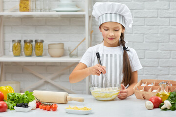 little child girl in cap and an apron kneads with steel whisk the dough in the kitchen. Whisking.