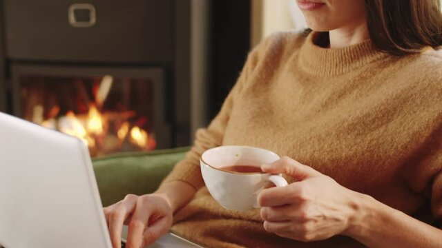 Close up shot of young woman typing on laptop and drinking tea while working remotely at home near fireplace
