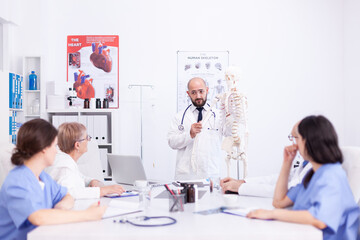 Fototapeta premium Expert radiologist demonstrating on skeleton during briefing with medical staff in hospital meeting room. Clinic therapist talking with colleagues about disease, medicine professional