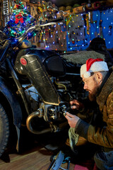 Obraz na płótnie Canvas A man in a santa claus hat repairs a motorcycle in the garage on New Year's Eve.