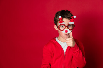 Pretty boy celebrating Christmas wearing funny Christmas glasses in front of red background