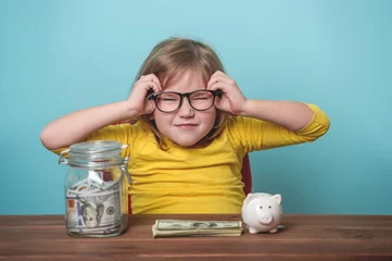 Fotobehang Funny child girl in glasses with saving money in jar and piggy bank on blue background. Buying eyewear opportunity. Save money on frames. vision problems. Negative emotion facial expression feelings © shara