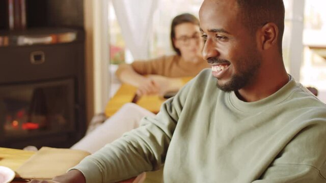 Close up shot of cheerful young Afro-American man messaging on laptop at home while his wife playing the guitar near fireplace in the background
