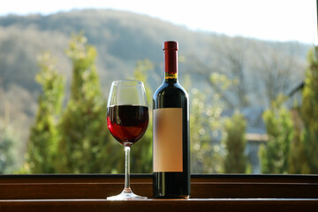 Fototapeta premium Glass and bottle of red wine on wooden windowsill against mountain background