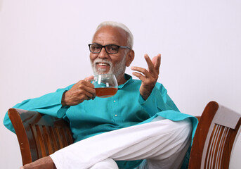 Indian old man having green tea while sitting on chair.