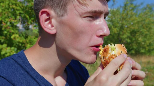 Young Man Having A Snack. A Guy Is Sitting On A Bench In The City Park And Biting A Burger