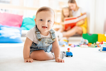Baby playing with toys while mother and daughter reading book on background, family time, education