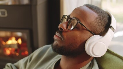 Close up shot of relaxed Afro-American man in wireless headphones leaning back in armchair with eyes closed and listening to music while sitting near fireplace at home - Powered by Adobe