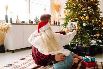 Christmas online holiday. Young couple having video call with their family or friends. Smiliтg couple in a santa hat uses a digital tablet near decorated festive tree at home. Virtual meeting. Covid